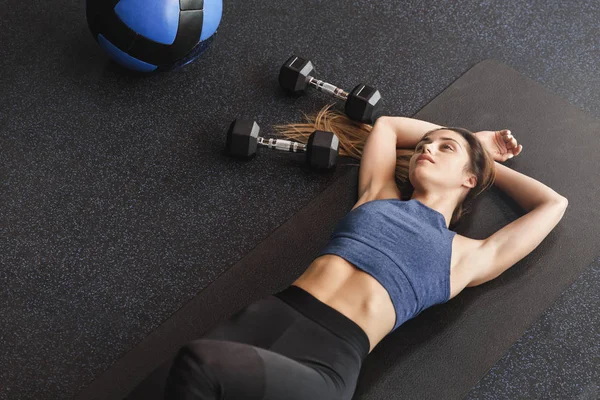 Upper angle dreamy sensual young sportswoman with six-pack, perfect body shape, lying on rubber mat at gym floor, thinking as rest before starting functional training with dumbbells and medicine ball — Stock Photo, Image