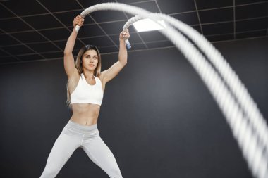 Eğitim, spor ve fonksiyonel eğitim konsepti. Motive olmuş güçlü ve formda bir bayan atlet, beyaz giysili bir kadın spor salonundaki savaş halatlarıyla egzersiz yapıyor.