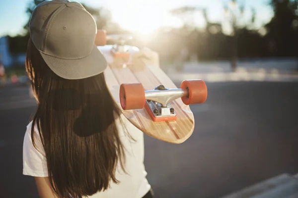 Nahaufnahme von hinten Skater Teenager Mädchen führen aktiven Lebensstil, tragen grauen Schnappschuss, tragen Holz Penny Board auf der Schulter, bewegen sich in Richtung Sonne auf Beton Skatepark, Stunts und Tricks ausführen — Stockfoto