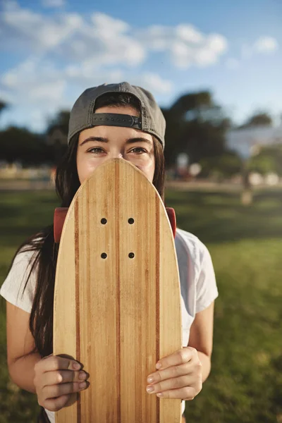 Vertikální záběr bezstarostný dospívající hipster dívka v šedé snapback, kryt tvář s dřevěnou penny palubě, oči s úsměvem a zírající fotoaparát, teenager těší letní dovolenou na skateboardu v parku — Stock fotografie