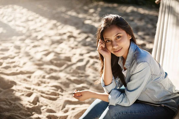Bara en mysig sommarkväll på stranden. Attraktiv ung leende kvinna i jeans jacka, jeans sitta bekväm hängmatta, fötter på sand glatt blickande kamera, romantisk dag under soliga solstrålar — Stockfoto