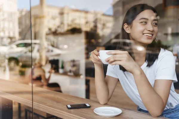 Freizeitkonzept. attraktive unbeschwerte junge asiatische Studentin Stop-Buy-Café in der Nähe der Universität, um köstlichen Cappuccino zu genießen, sitzen in der Nähe von Fenster schlanken Tisch, in der Nähe liegen Smartphone, im Gespräch mit Freund — Stockfoto