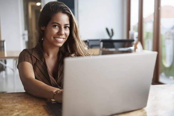 Atractiva y exitosa mujer hispana sonriendo mientras toma el teclado de las manos, usando el portátil, mira la cámara feliz, trabaja a distancia, freelancer escribe un nuevo post. Estudio femenino en el espacio de co-trabajo, beber café — Foto de Stock