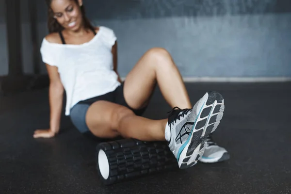 Woman sitting on gym floor in activewear, white t-shirt, face blurred as focus on legs doing exercise with functional training equipment, foam roller, smiling satisfied, motivated get perfect body