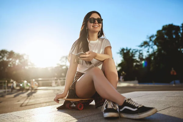 Glück, Freizeit und urbaner Lebensstil. Unbekümmerte Teenagerin mit Sonnenbrille übt bei sonnigem Sommertag im Skatepark Skateboarding-Tricks, Smartphone in der Hand — Stockfoto