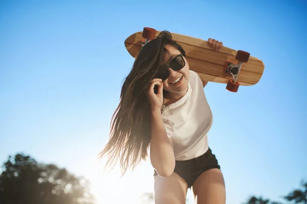 Спорт, міський спосіб життя та концепція молоді. Весела дівчина нахиляється, щоб виглядати камерою, тримає дерев'яну дошку на плечі, торкається сонцезахисних окулярів, насолоджується сонячними літніми канікулами, катаючись на скейтборді — стокове фото