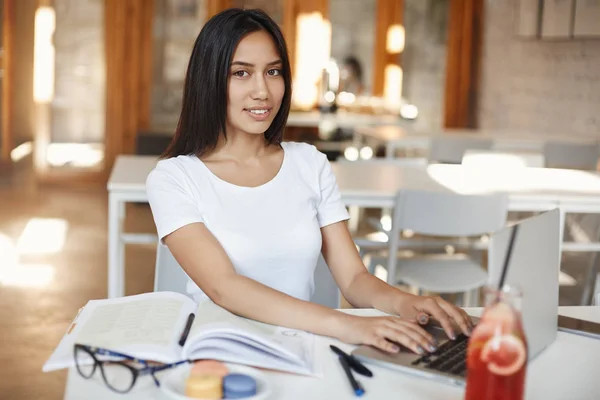 Atractiva mujer sonriente freelancing, reunir material en Internet, escribir en el teclado del ordenador portátil y la cámara sonriente, estudiante alegre trabajando, idiomas de estudio con el sitio de educación de la cafetería cerca del campus — Foto de Stock