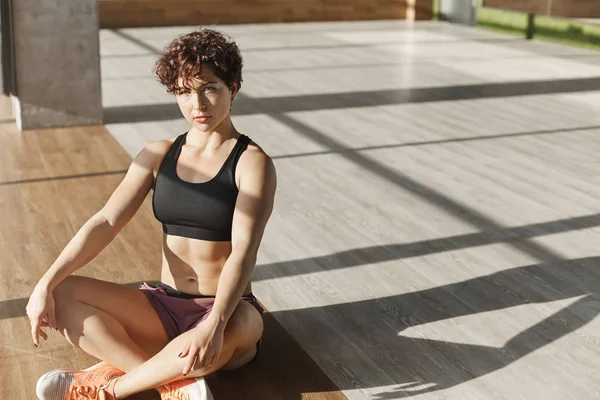 Motivation, santé et concept sportif. Prise de vue angle supérieur, jolies sportives avec coupe courte bouclée en sportsbra, short, pose de méditation de lotus assis, caméra de recherche déterminée dans la salle de gym — Photo