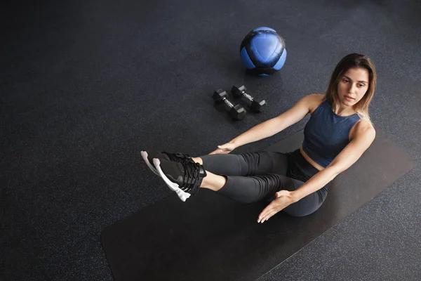 Upper-angle shot motivated young female athlete, sportswoman workout in gym, lying rubber mat near medicine ball, two dumbbells, functional training equipment, perform crunches with lifting legs