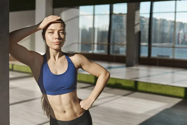 Athletic and strong good-looking young fitness instructor, sportswomen in sportsbra, showing six-pack perfect body, leaning on wall in gym, dance studio, mirrors behind, looking camera confident — 스톡 사진