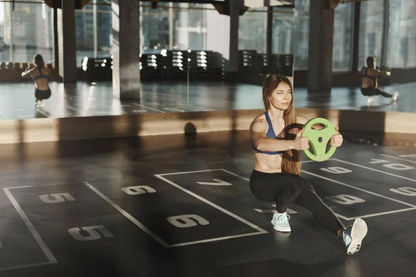 Woman in middle of functional training in gym. Attractive caucasian athletic sportswoman do squats with extended foot, holding medicine ball and lookig forward at window, workout alone — 스톡 사진
