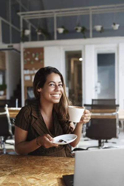 Verticale schot prachtige Spaanse vrouw neemt koffiepauze, zitten alleen co-working ruimte of cafe, laptop geopend op tafel, ze houdt kopje als smaakt heerlijke cappuccino, glimlachende camera gelukkig — Stockfoto