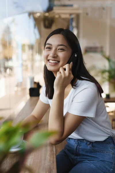 Vertikale Aufnahme lächelnd, Millennial süße asiatische Mädchen im Café, sprechen per Smartphone, halten Handy in der Nähe Ohr, warten auf Freund, Kaffee trinken, betrachten draußen als Blick durch das Fenster — Stockfoto
