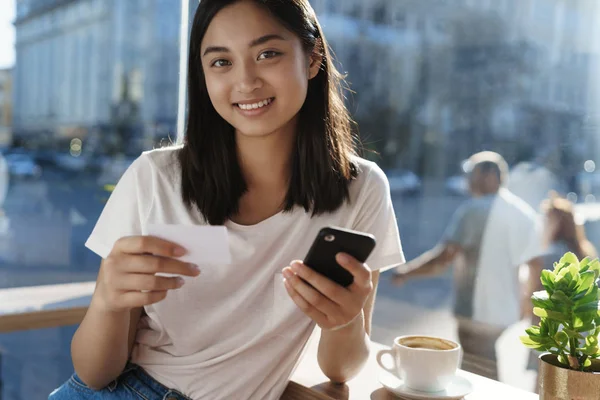 Gaya hidup perkotaan, kota, konsep masyarakat. Lovely tersenyum gadis asia dalam kaos putih kasual, memegang smartphone dan kartu kredit, duduk di dekat jendela kafe di meja kopi, di belakang orang yang lewat jalan berjalan — Stok Foto