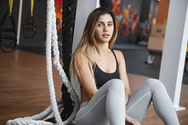 Sport, healthy lifestyle concept. Strong attractive sportswoman, female athlete in sportsbra, grey leggings sit near battle ropes and functional training equipment, catching breath after workout