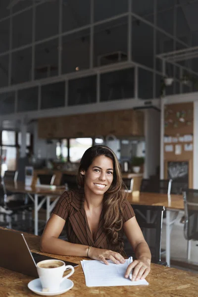 Pengambilan gambar vertikal wanita pengusaha brazilian muda yang sukses, pengusaha duduk di kafe, bekerja sama dengan laptop terbuka, minum cappuccino dan menulis catatan di atas kertas, tersenyum — Stok Foto
