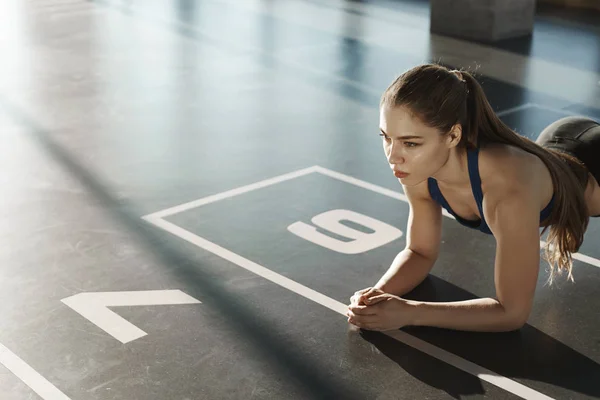 Endurance, sport and healthy lifestyle concept. Side-shot focused young athletic woman, standing in plank on gym floor, looking forward, workout, training to stay fit, morning fitness exercises — 스톡 사진