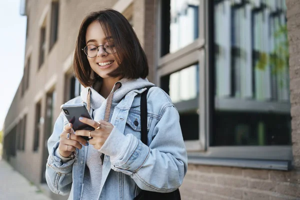 Молодая педик, студентка колледжа, стоящая на улице, заказывающая такси с помощью приложения на смартфоне, улыбающаяся как друг, встречающаяся с командой за пределами кампуса, прокручивающая лента новостей в социальных сетях — стоковое фото