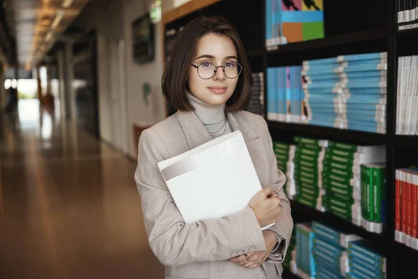 Berhasil wanita muda terdaftar di universitas, belajar keras mendapatkan gelar, bekerja pada proyek, memegang dokumen dan tersenyum kamera dengan santai ekspresi, berdiri di dekat tumpukan buku di aula — Stok Foto