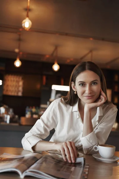 Concepto de negocios y mujeres. Retrato vertical de una hermosa dama sentada en la cafetería, inclinada mesa de café y leyendo la revista de moda mientras bebe cappuccino, sonriendo tiernamente cámara — Foto de Stock