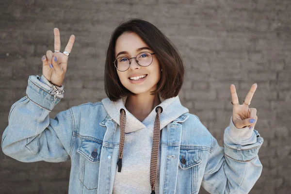 รูปภาพใกล้ชิดของสาวน่ารักในแว่นตา นักเรียนมหาวิทยาลัยแสดงท่าทางสันติภาพคาวาย และกล้องยิ้ม สนุกกับเยาวชน ชีวิตที่ปราศจากมหาวิทยาลัย ยืนอยู่ใกล้กําแพงอิฐบนถนน — ภาพถ่ายสต็อก