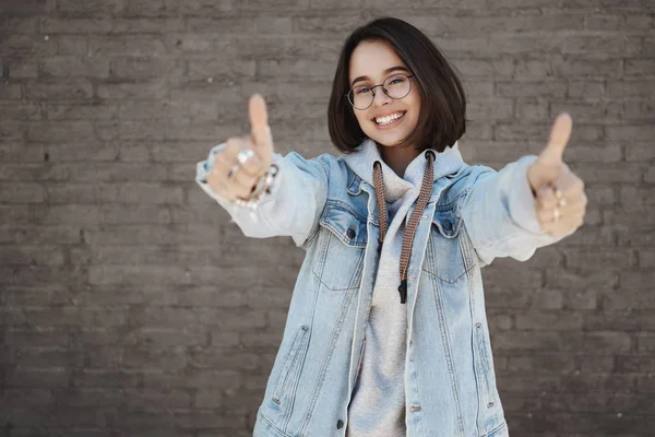 Super cool offer. Smiling carefree and happy young positive girl in glasses with short hair, wear denim jacket, show thumbs up, encourage join awesome company, language center, recommend product