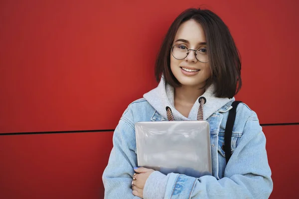 Close-up ao ar livre retrato de estudante europeu feminino em óculos, menina atraente de pé perto da parede vermelha, segurando laptop em mãos, câmera sorridente com expressão relaxada alegre — Fotografia de Stock