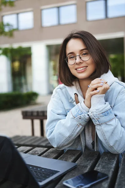 Leren, onderwijs en jeugdconcept. Verticaal portret van meisje zittend op bank buiten in park met laptop en smartphone, glimlachende camera, studeren op frisse lucht, lokale wifi aansluiten — Stockfoto