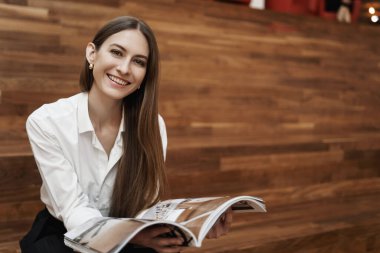 Güçlü kadın ve iş kavramı. Genç beyaz kızı merdivenlerde oturtup, yemek molası veriyor, moda veya tasarım dergisi okuyor, gülümsüyor, çalışma alanında rahatlıyor.