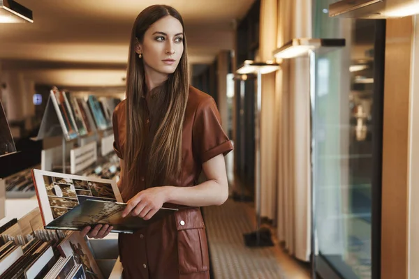 Elegant young alluring woman shopping at vintage bookstore, holding magazine, turn around look outside window with thoughtful sensual expression, picking new books to read, explore design