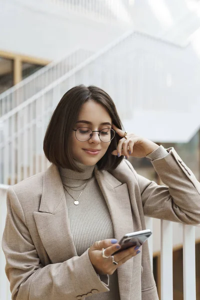 Vertikales Porträt einer gut aussehenden jungen stilvollen Unternehmerin, die eine neue Karriere beginnt, in der Büro-Lobby auf ihr Interview wartet, ihr Handy in der Hand hält, Freundin SMS schreibt und auf dem Bildschirm lächelt — Stockfoto