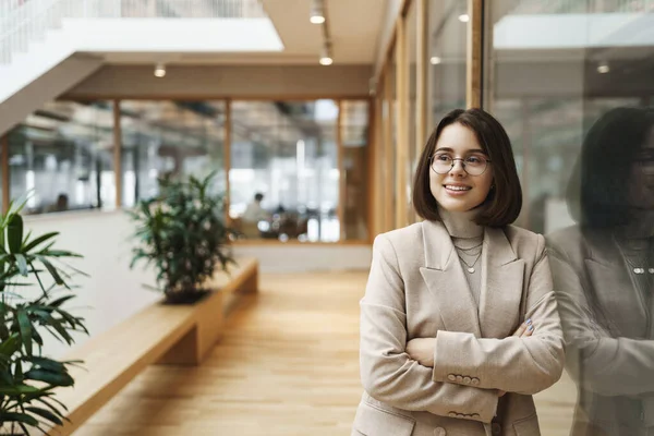 Business, education and women concept. Portrait of successful young caucasian woman in elegant jacket, leaning on glass wall in office hall waiting for coworker business center, smiling happy — Stock Photo, Image
