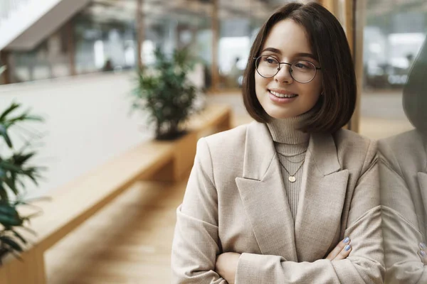 Primo piano ritratto di successo e fiducioso giovane donna iniziare la carriera, guardare determinato a trovare lavoro, in attesa di intervista, magra sulla parete di vetro in business center, sorridente e guardando lontano soddisfatto — Foto Stock