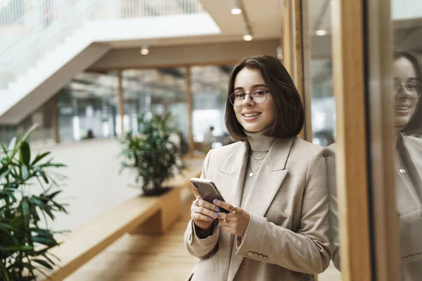 Portrait of successful happy young woman start prominent career, sending message, use mobile phone and smiling at camera as waiting for someone in office hall, reception, meeting business partners — Stock Photo, Image