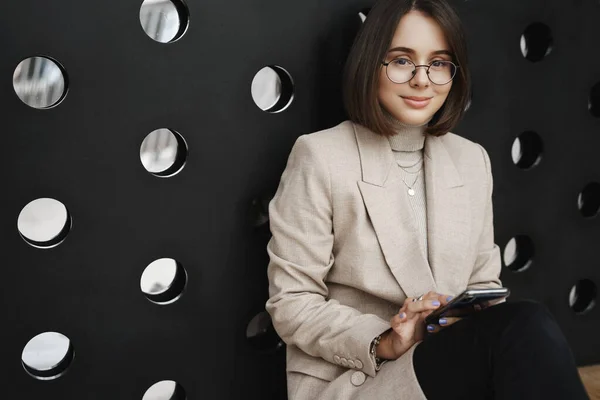 Retrato de mujer joven y elegante feliz en gafas y chaqueta, sentado en el suelo y la pared magra, sonriendo a la cámara como mensajería en el teléfono móvil, desplazamiento de alimentación de Internet, amigo mensajes de texto mientras espera — Foto de Stock