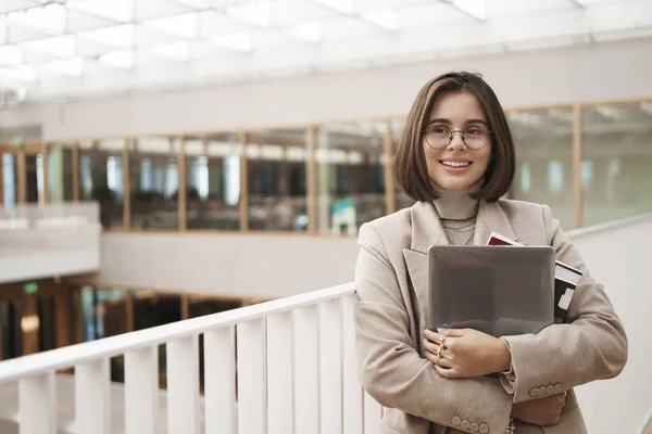 Education, career and university concept. Portrait of happy young pretty girl in jacket and glasses, holding laptop with worksheets and books, leaning on staircase and gazing away cheerful — Stock Photo, Image