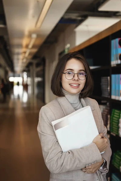 Potret vertikal gadis Kaukasia yang cantik dengan jaket yang berdiri di perpustakaan universitas dengan seprai kerja dan buku, senyum bahagia berpaling memimpikan, belajar untuk proyek atau kursus, berpose di aula — Stok Foto
