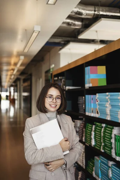 Ritratto verticale di giovane elegante studentessa felice in piedi nella sala del campus o biblioteca vicino alla libreria, tenendo libro e fogli di lavoro macchina fotografica sorridente, studiando per le classi, preparare il rapporto — Foto Stock