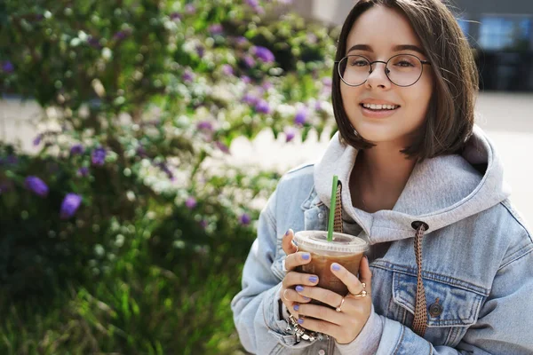 Detailní portrét šťastné atraktivní mladé ženy s krátkým účesem v brýlích a džínové bundě, držící odnášející ledovou kávu, užívající si jarního slunečného dne ve městě, usmívající se kamera — Stock fotografie