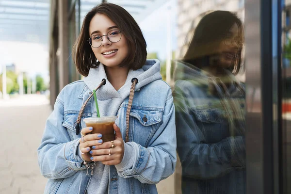 Gadis muda yang menarik dalam kacamata, jaket denim, berjalan santai di kota, menikmati akhir pekan, minum es latte, bersandar pada dinding bangunan dan tersenyum kamera dengan ekspresi santai bahagia — Stok Foto