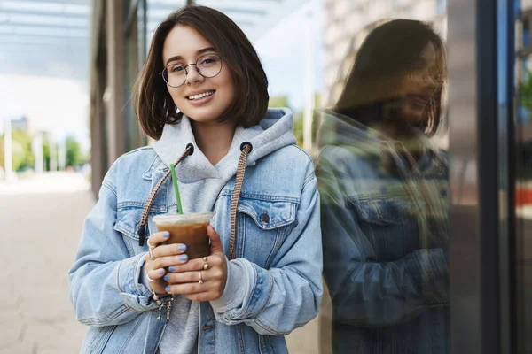 Mladá atraktivní dívka v brýlích, džínová bunda, neformální procházka po městě, užívání si víkendů, popíjení ledového latté, opírání se o zeď budovy a usmívání fotoaparátu se šťastným uvolněným výrazem — Stock fotografie