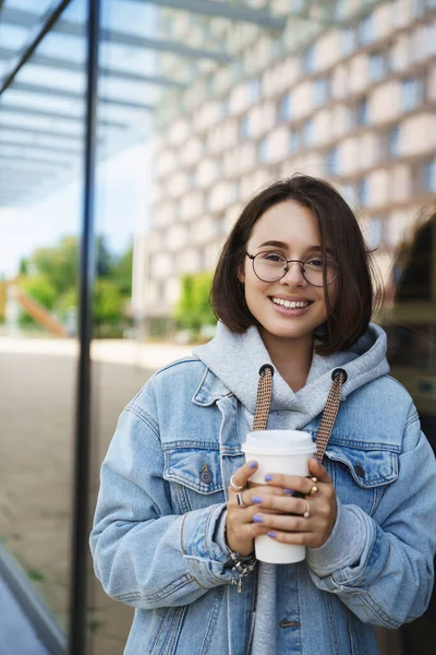 Vertikální portrét šťastně se usmívající mladé přítelkyně s krátkými vlasy, v džínové bundě a brýlích, držící odnášející kávu, kráčející s kamarádkou a smějící se přes vtipnou konverzaci — Stock fotografie