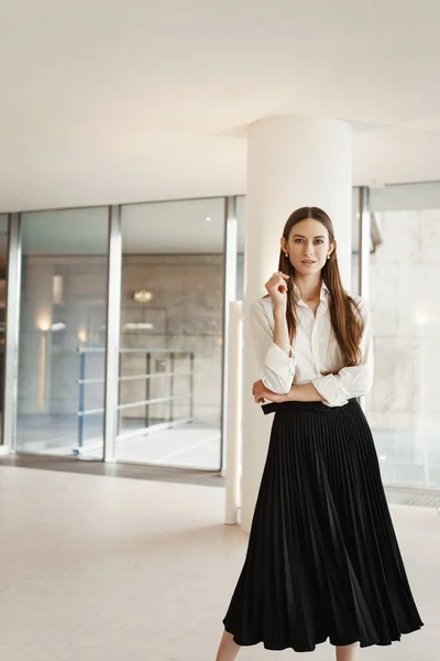 Vertical portrait of independent strong woman in black formal skirt and white blouse, working in office, standing confident in white corridor, expressing self-assurance and success