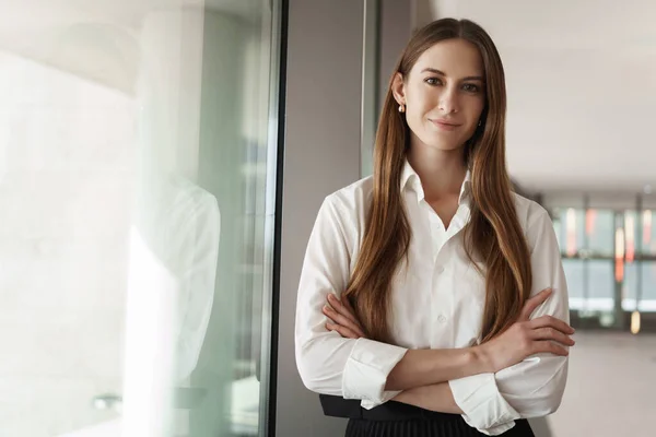 Pleasant young female assistant, manager standing in office corridor near window, with pleased confident expression, cross hands over chest showing readiness, business and women concept — Stock Photo, Image