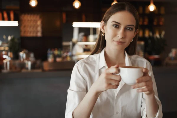 Kopi pertama. Wanita muda yang percaya diri dengan blus putih, memegang cangkir cappuccino, duduk di kafe dengan kolega setelah bekerja keras, makan siang istirahat, menikmati rasa minuman yang enak — Stok Foto