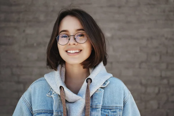 การศึกษา อาชีพสําหรับคนหนุ่มสาว รูปภาพใกล้ชิดของสาววัย 20 ที่ยิ้มอย่างมีความสุขในแว่นตาและแจ็คเก็ตเดนนิมเหนือเสื้อฮู้ด ดูกล้องที่พอใจและมีความสุข ยืนอยู่ข้างนอกเหนือกําแพงอิฐ — ภาพถ่ายสต็อก