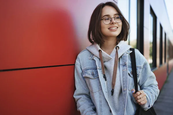 Kesempatan karir, pendidikan dan konsep freelance. Happy santai gadis yang cukup aneh dalam kacamata, jaket denim, memegang tas tote, dinding bangunan merah ramping dan berpaling senang, merenungkan musim semi — Stok Foto