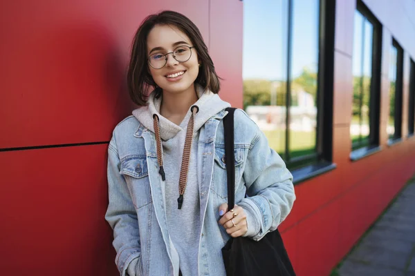 Primo piano ritratto di allegra bella giovane 20s donna in occhiali, studente o dipendente, fotocamera sorridente felice, portando tote bag, magra muro di mattoni rossi del campus, godendo il tempo libero — Foto Stock