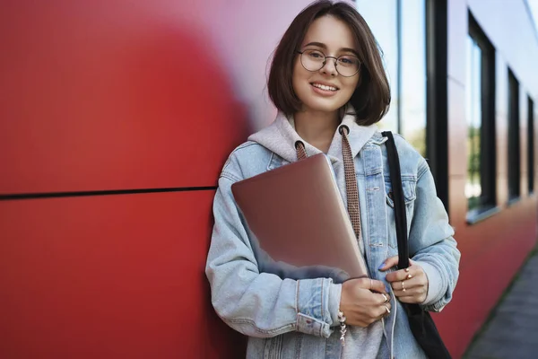 Ritratto in vita di giovane programmatrice, IT o smm manager, vuole lavorare all'aperto nel parco durante il bel tempo primaverile, in piedi con il computer portatile magra muro di mattoni rossi, sorridente fotocamera felice — Foto Stock