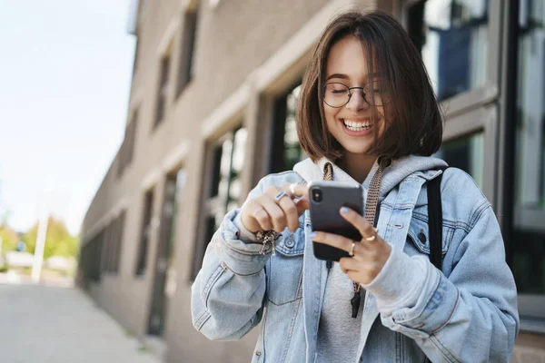 Youth, technology and spring concept. Cheerful pretty young woman in glasses, denim jacket, laughing and smiling looking mobile phone display, send funny message, texting as standing on street — Stock Photo, Image